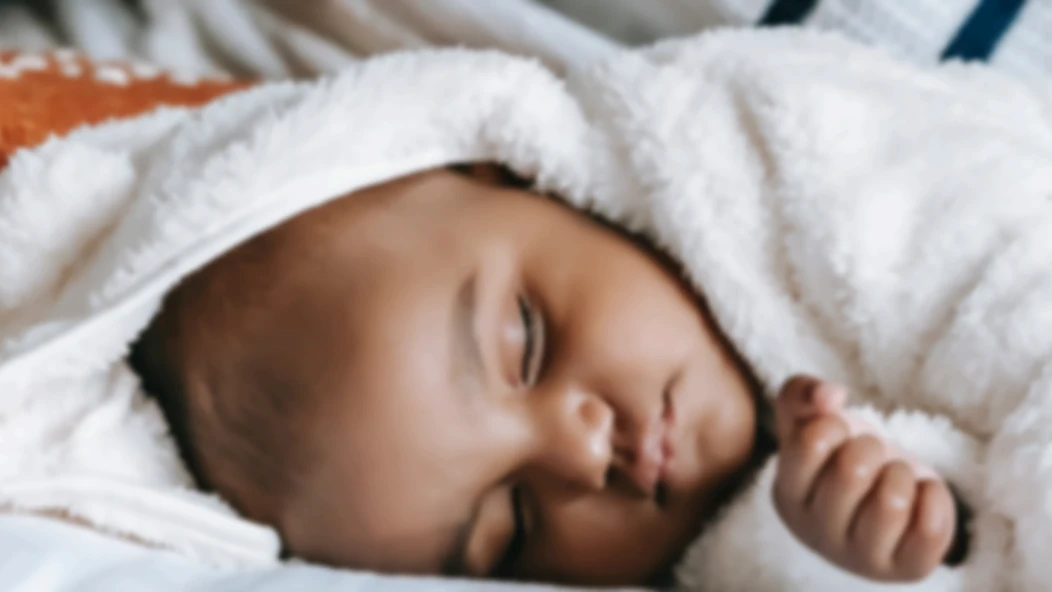A close-up photograph of a sleeping infant wrapped in a soft, white textured blanket.