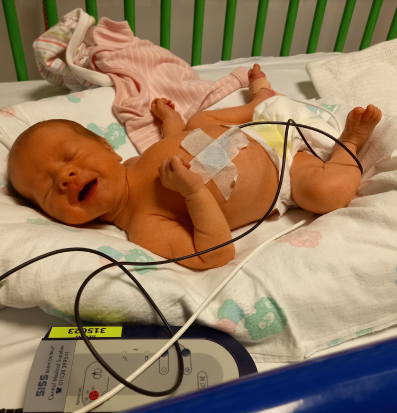 Newborn baby lying on its back in a hospital crib, connected to medical monitoring equipment, with a sensor on its chest and wires leading to a monitor. The baby appears to be crying, and there are baby clothes nearby. The environment suggests a hospital setting with a focus on infant care.