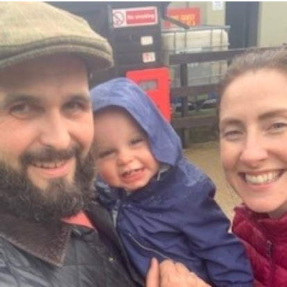 A family selfie featuring two adults and a young child outdoors. The man on the left, wearing a flat cap and beard, is smiling at the camera. The child in the middle is wearing a blue hooded jacket and has a joyful expression. The woman on the right, with a red jacket and smiling, is looking at the camera. They seem to be standing in front of a farm or industrial setting, as evidenced by the equipment in the background.